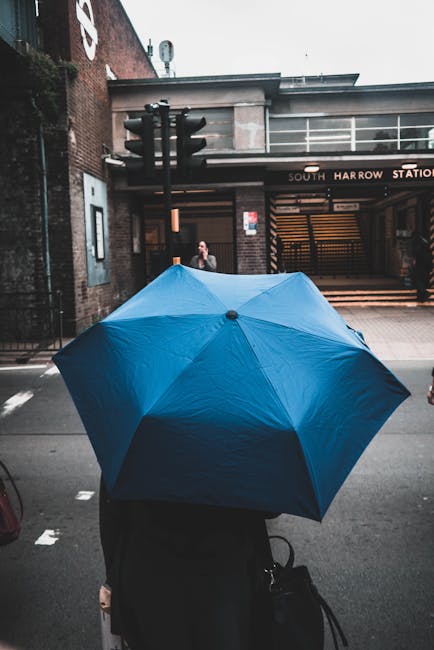 A person holding a large, vibrant blue umbrella stands on a city street in front of South Harrow Station entrance. The individual is dressed in dark clothing and carries a black bag. The station build