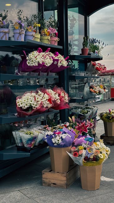 A display of fresh floral arrangements on metal shelves outside a flower shop, featuring bouquets wrapped in colorful paper and ribbon. The arrangements include white chrysanthemums, pink lilies, purp