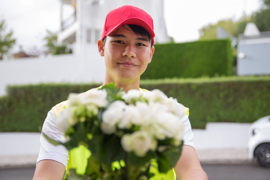 A delivery person wearing a red cap and a high-visibility yellow vest holds out a bouquet of fresh white roses and lilies, wrapped in clear cellophane, towards the camera during a flower delivery. The