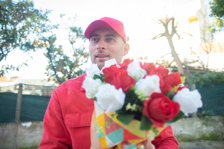 A man dressed in a red jacket and matching red cap is holding a floral bouquet in front of him, featuring red roses, white flowers, and green foliage wrapped in colorful paper. The background shows a 