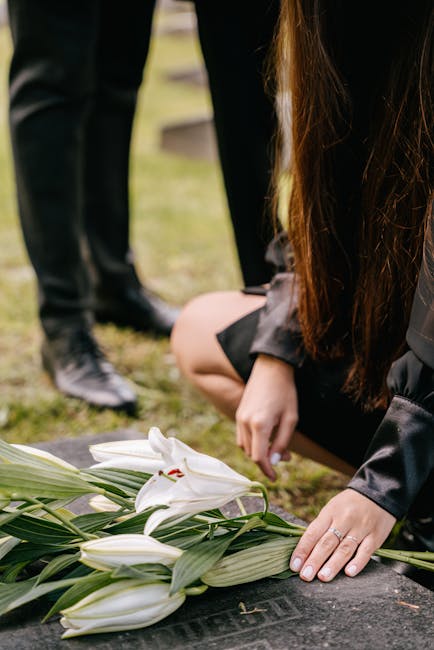 A person with long dark hair, dressed in black, is kneeling at a graveside, carefully arranging a floral tribute consisting of white lilies and green foliage. The lilies are fresh, with delicate petal