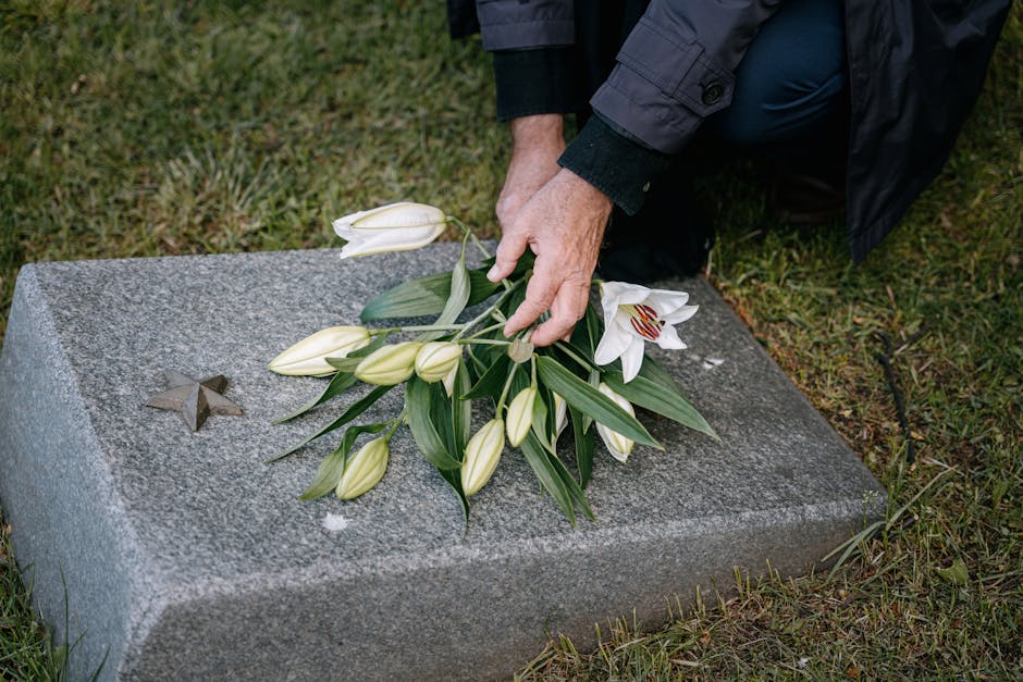 A person dressed in dark clothing, kneeling on grass, arranging a bouquet of fresh white lilies with large green leaves on a grey granite headstone at a memorial site. The bouquet includes several clo