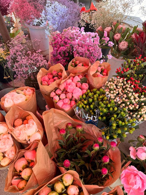 A vibrant display of freshly cut flowers arranged in brown paper wrappings, featuring pink peonies, green berries, and various shades of pink and white blossoms. The flowers are placed on a table at a
