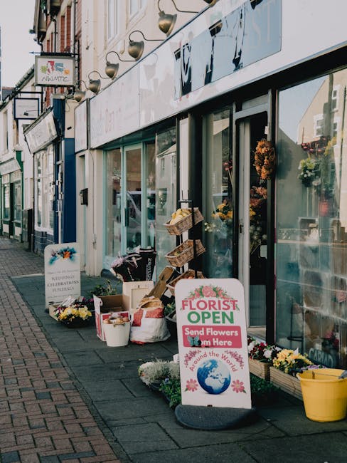 A storefront of Florist Harrow displaying a variety of fresh flowers arranged in baskets and buckets on the sidewalk outside the shop. The floral display includes colorful blooms such as yellow, white