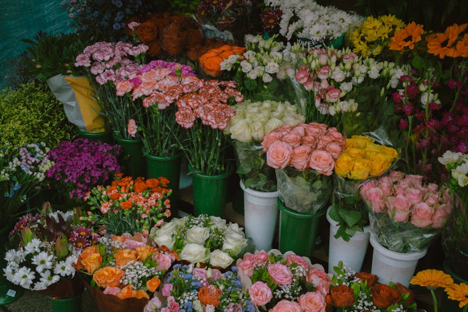 A vibrant display of fresh floral arrangements and bouquets arranged in white and green plastic containers on a flower shop shelf. The flowers include pink, peach, yellow, and white roses, intersperse