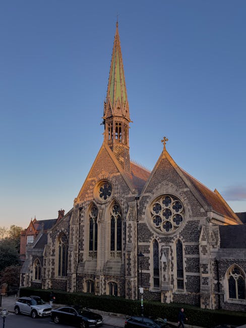 A historic stone church with a tall, pointed spire topped with a cross, featuring intricate Gothic-style windows with decorative tracery and circular motifs. The church's stone exterior showcases a mi