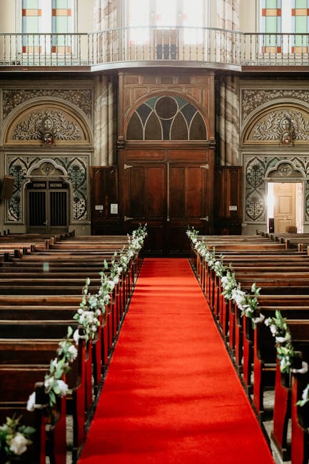 An indoor church aisle set for a wedding service, featuring a vibrant red carpet extending from the entrance towards the altar. On either side of the aisle, wooden pews are decorated with elegant flor